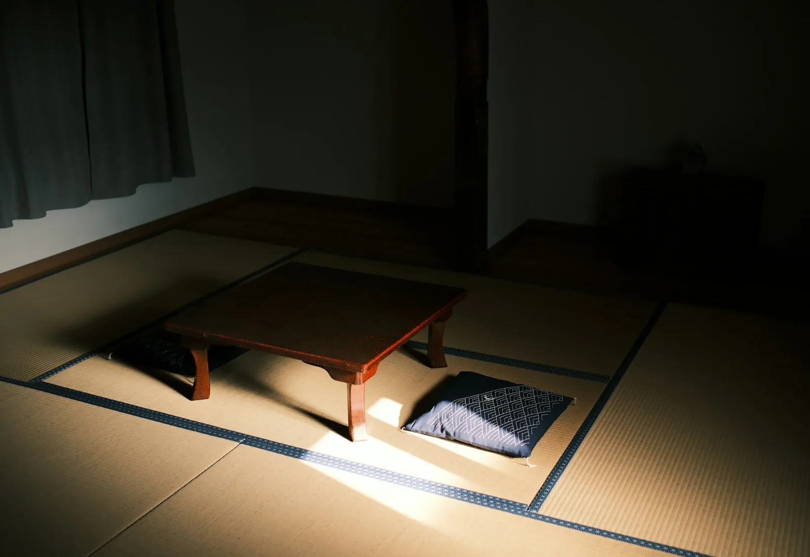 畳の座敷に置かれた朱の座卓と座布団、そこへ落ちる一条の光