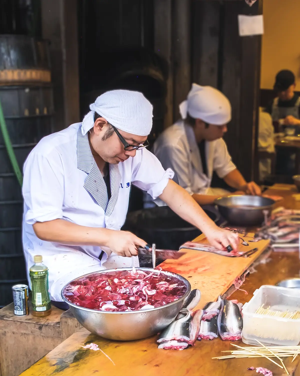 白い手ぬぐいをかぶった料理人が、まな板の上で魚を包丁でおろしているところ