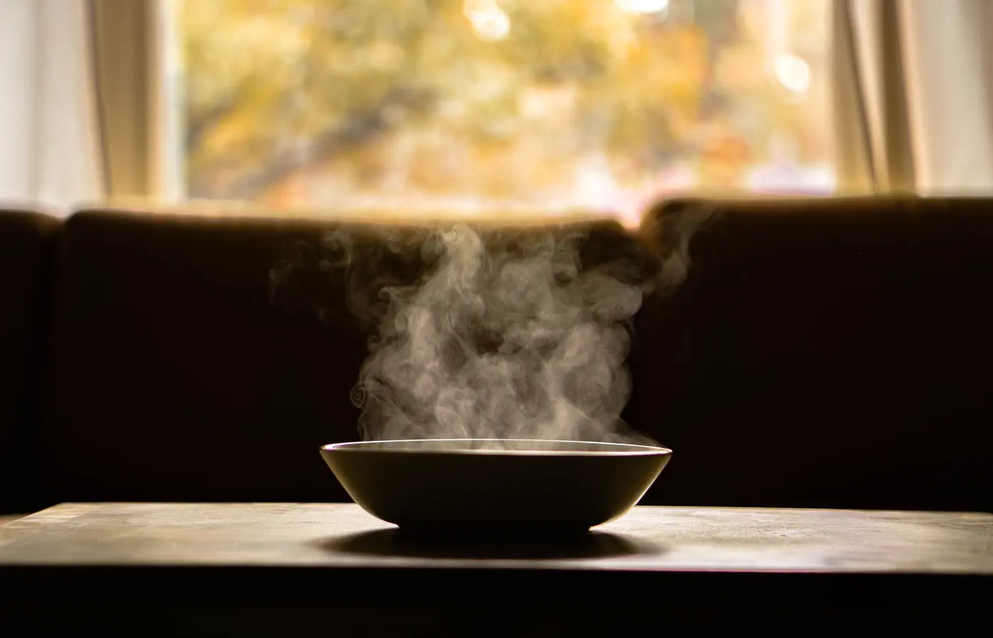 窓のあかりを背にした卓の上で、椀から湯気が立ちのぼっているところ